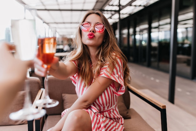 Maravilhosa jovem modelo feminino bebendo coquetel no restaurante. Foto interna de uma garota romântica em vestido listrado, posando com beijando a expressão do rosto no café.