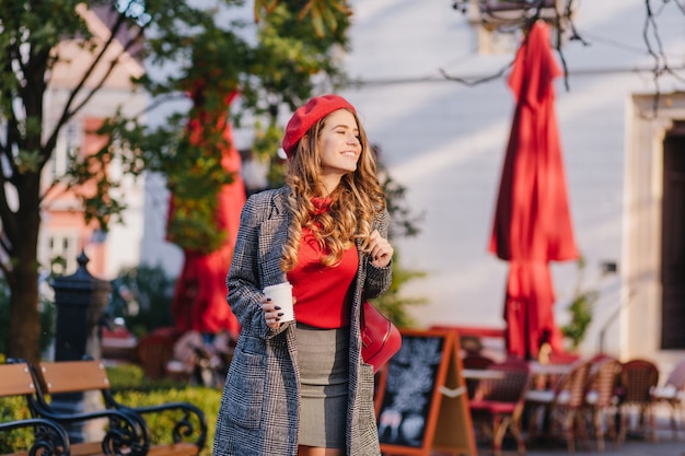 Maravilhosa modelo feminina com roupas cinza, andando na rua com uma xícara de café