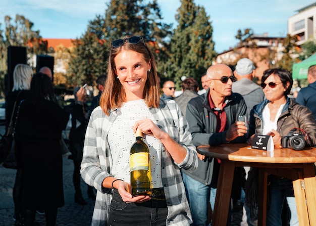 Maribor, eslovênia - 11 de outubro de 2019: jovem sorridente usando óculos escuros segura garrafa de vinho estilo casual de mulher. pessoa do sexo feminino se divertindo. boa celebração do feriado. retrato de estilo de vida ao ar livre.