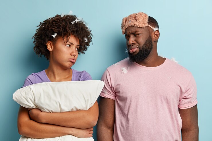 Foto grátis marido e mulher de pele escura chateados e aborrecidos olham um para o outro com expressões faciais tristes e sombrias, têm mau humor depois de dormir por um curto período de tempo, usa máscara de dormir, isolado sobre a parede azul.