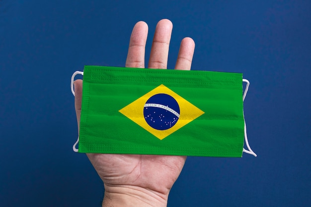 Máscara cirúrgica sobre fundo azul com bandeira do brasil - mão de homem segurando