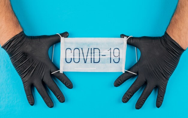 Máscara médica com texto covid-19 nas mãos com vista superior de luvas pretas