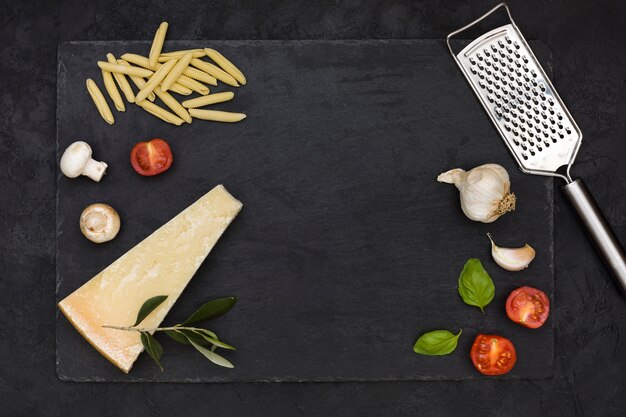 Massa garganelli italiano cru com ingredientes e ralador de queijo na ardósia rocha contra o pano de fundo preto