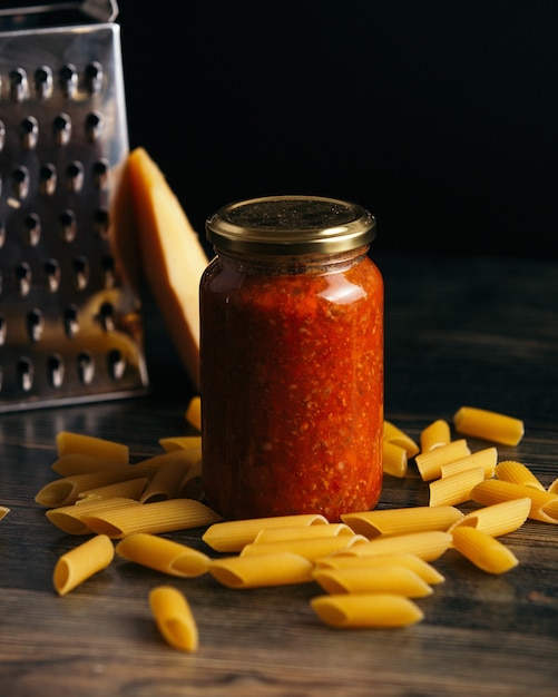 Massa penne e um pote de molho na mesa com queijo e um ralador no fundo