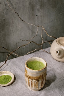 Matcha de chá verde quente japonês tradicional espumoso em xícara de cerâmica artesanal, matcha em pó, bule de chá em pé sobre toalha de mesa de linho cinza. bebida saudavel