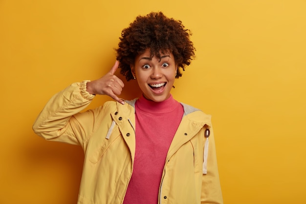 Me ligue de volta. Mulher simpática e positiva com cabelo encaracolado, gesticula perto da orelha, mostra gestos no telefone, parece alegre, tenta entrar em contato com amigos ou parentes, fica em pé sobre uma parede amarela