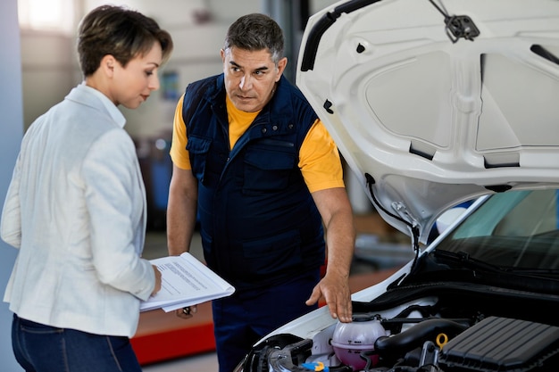 Mecânico de automóveis e gerente feminina falando enquanto examinava avaria do motor do carro na oficina de reparação automóvel