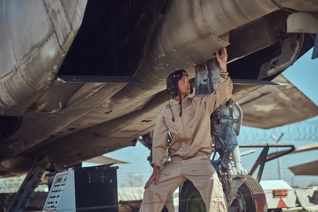 Mecânico de uniforme e capacete de voo, realiza a manutenção de um caça-interceptador de guerra em um museu a céu aberto.
