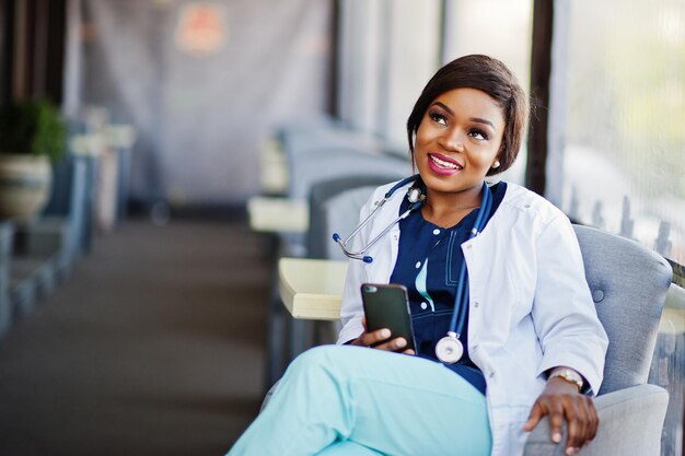 Médica afro-americana com estetoscópio olhou para o telefone e sentada na clínica