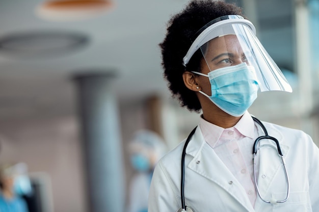 Médica afro-americana pensativa com escudo facial e máscara no hospital