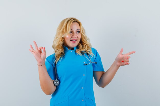 Médica apontando de lado enquanto mostra um gesto ok com uniforme azul e parece feliz.