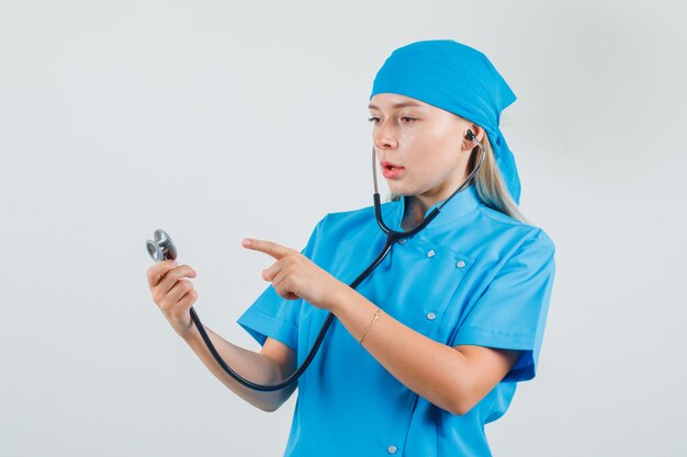 Médica, apontando o dedo para o estetoscópio em uniforme azul e olhando séria.