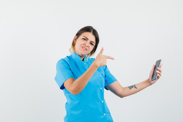 Médica apontando para o celular com uniforme azul e parecendo confiante