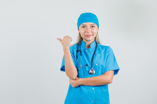 Médica, apontando para o lado com o polegar em uniforme azul e parecendo alegre.