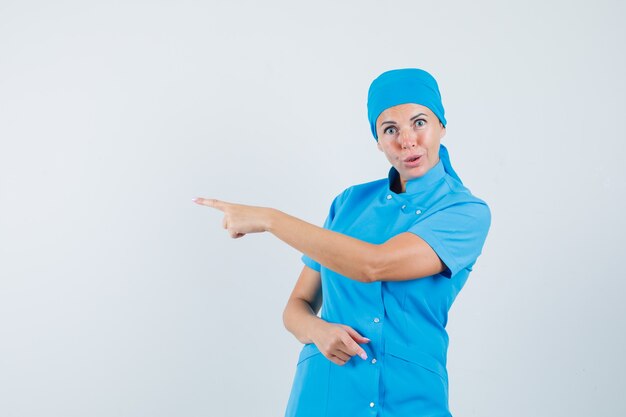Médica apontando para o lado com uniforme azul e parecendo hesitante. vista frontal.