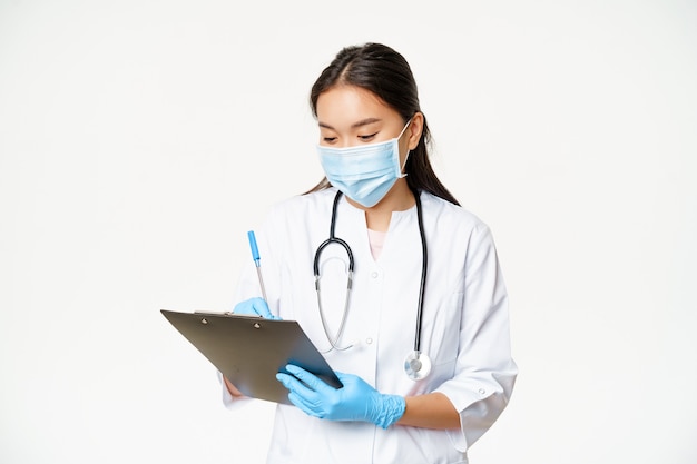 Médica asiática, escrevendo as informações do paciente na área de transferência, prescrever ou diagnosticar, de pé na máscara médica com luvas de borracha, fundo branco.