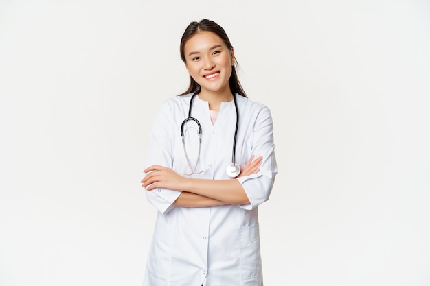 Médica asiática, médica em uniforme médico com estetoscópio, braços cruzados no peito, sorrindo e parecendo profissional, com fundo branco