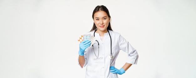 Médica asiática mostrando medicação de pílulas segurando vitaminas e usando luvas de borracha sobre fundo branco
