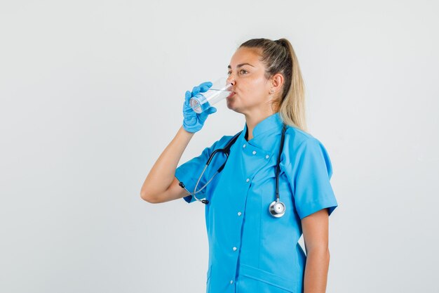 Médica bebendo água em uniforme azul, luvas e parecendo com sede.