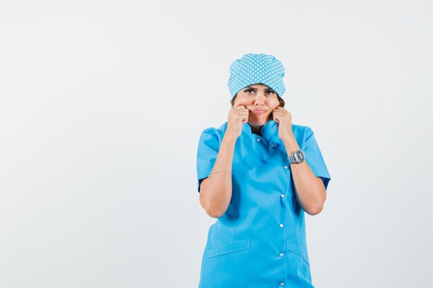 Médica beliscando as bochechas com uniforme azul e parecendo triste