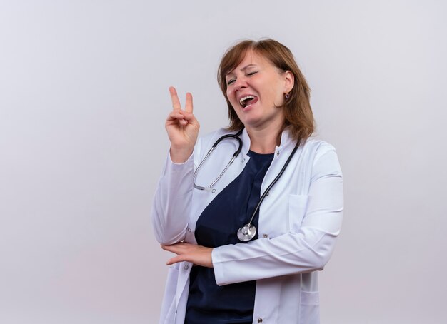 Médica brincalhona de meia-idade usando túnica médica e estetoscópio, fazendo o sinal da paz e mostrando a língua na parede branca isolada com espaço de cópia