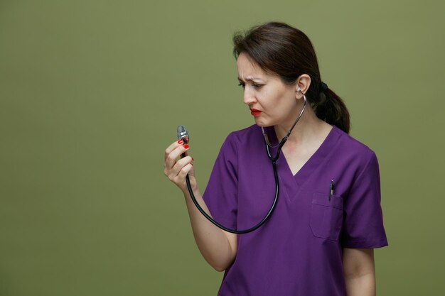 Médica carrancuda de meia-idade vestindo uniforme e estetoscópio agarrando estetoscópio olhando para ele isolado em fundo verde-oliva
