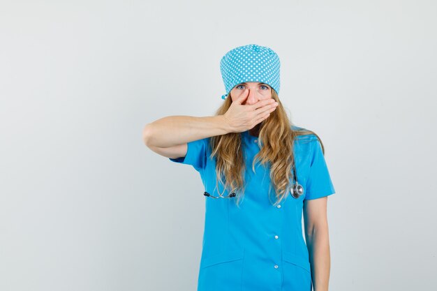Médica cobrindo a boca com a mão em uniforme azul e parecendo chocada.