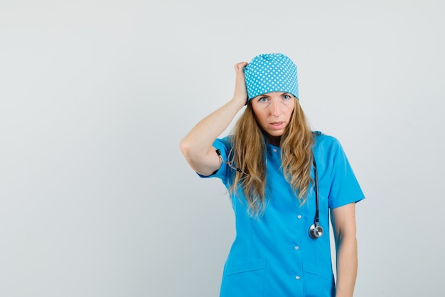 Médica coçando a cabeça em uniforme azul e olhando pensativa.