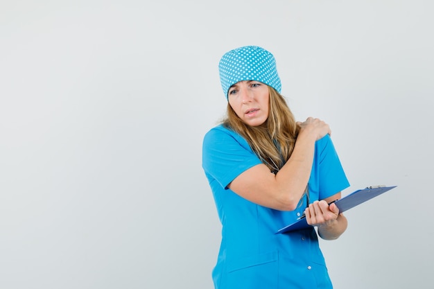 Médica com dor no ombro, segurando a prancheta em uniforme azul e parecendo cansada.