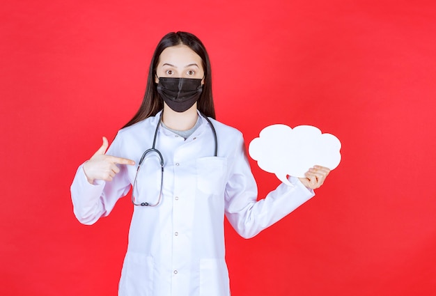 Foto grátis médica com estetoscópio e máscara preta, segurando uma mesa de informações em branco de forma de nuvem.