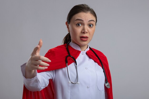 Médica com estetoscópio em uniforme médico branco e capa de super-herói vermelha apontando com o dedo indicador para a câmera, parecendo confusa em pé sobre uma parede branca