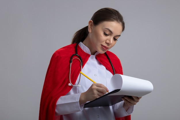 Médica com estetoscópio em uniforme médico branco e capa de super-herói vermelha segurando uma prancheta com páginas em branco escrevendo algo com uma cara séria em pé sobre uma parede branca