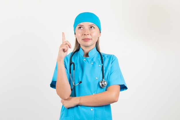 Médica com uniforme azul apontando o dedo para cima e sorrindo