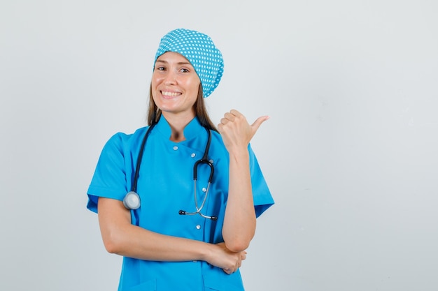 Médica com uniforme azul apontando o polegar para o lado e parecendo feliz