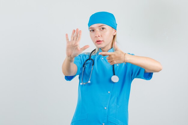 Médica com uniforme azul apontando para a palma da mão levantada e olhando séria