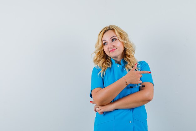 Médica com uniforme azul apontando para o lado e olhando para o espaço para texto