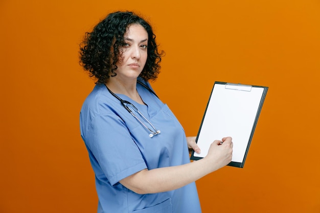 Médica confiante de meia-idade vestindo uniforme e estetoscópio em volta do pescoço em pé na vista de perfil segurando a área de transferência escrevendo nele com caneta olhando para câmera isolada em fundo laranja