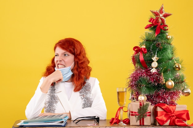 Médica de frente ao redor da árvore de natal e presentes sentada com uma máscara estéril