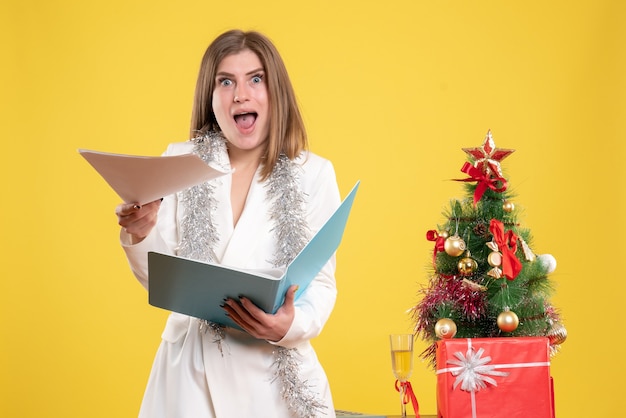 Médica de frente, de pé, segurando documentos na mesa amarela com uma árvore de natal e caixas de presente