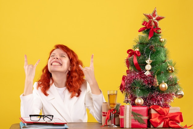 Médica de frente sentada atrás da mesa com os presentes de Natal e a árvore na mesa amarela