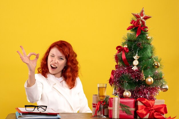 Médica de frente, sentada atrás da mesa, com os presentes de Natal e a árvore no fundo amarelo