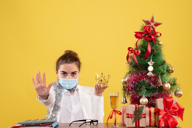 Médica de frente sentada com máscara estéril segurando uma coroa sobre fundo amarelo com árvore de Natal e caixas de presente