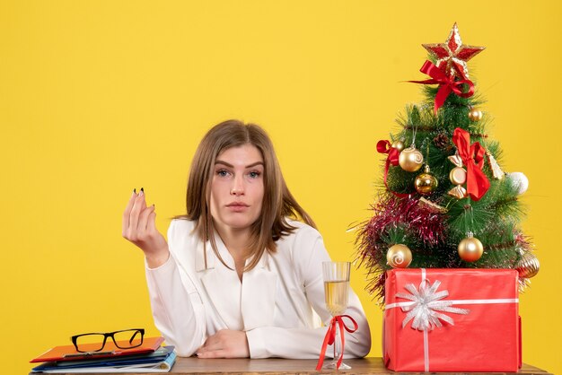Médica de frente sentada em frente à mesa na mesa amarela com árvore de Natal e caixas de presente