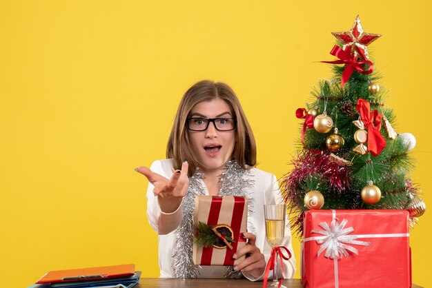 Médica de frente, sentada em frente à mesa, segurando um presente sobre fundo amarelo com árvore de Natal e caixas de presente