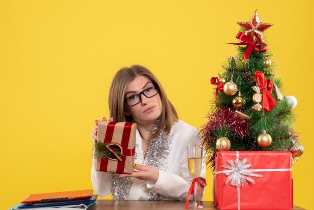 Médica de frente, sentada em frente à mesa, segurando um presente sobre fundo amarelo com árvore de Natal e caixas de presente