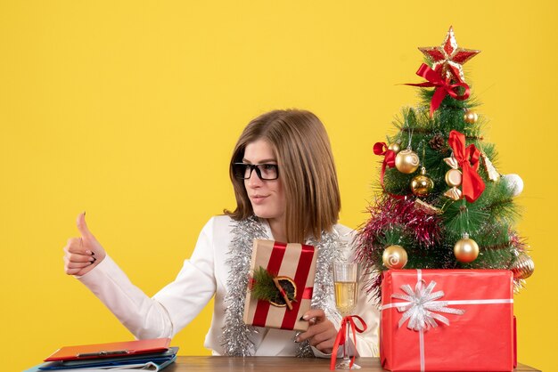 Médica de frente, sentada em frente à mesa, segurando um presente sobre fundo amarelo com árvore de Natal e caixas de presente