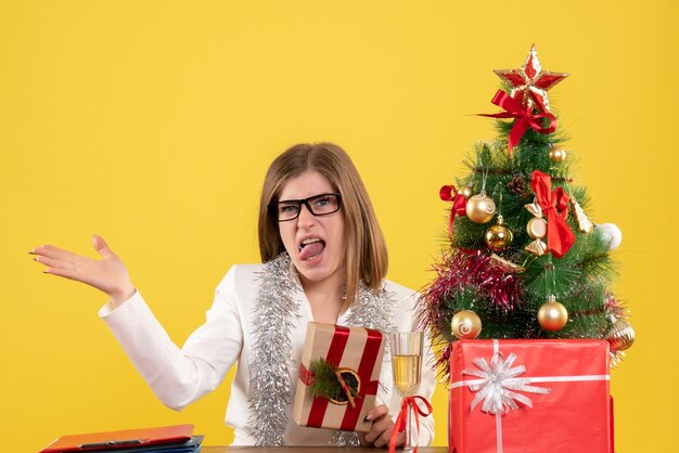 Médica de frente, sentada em frente à mesa, segurando um presente sobre fundo amarelo com árvore de Natal e caixas de presente