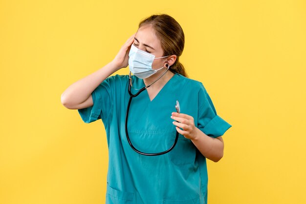 Médica de frente, tendo dor de cabeça na máscara sobre fundo amarelo, vírus da saúde pandêmico covid