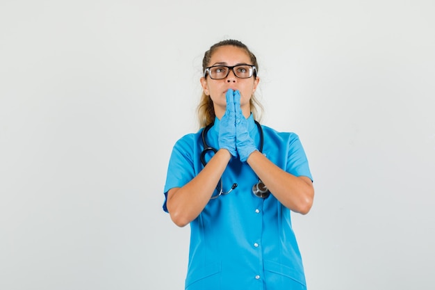 Médica de mãos dadas em gesto de oração em uniforme azul, luvas, óculos e parecendo esperançosa