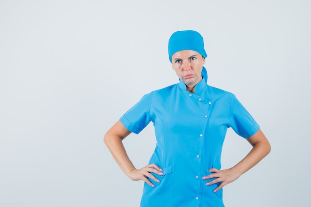 Foto grátis médica de mãos dadas na cintura em uniforme azul e olhando séria, vista frontal.